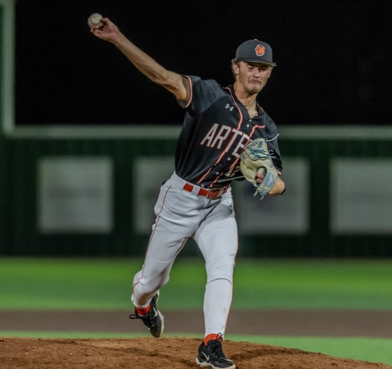 A teen boy in a black, orange and white baseball uniform pitches from the mound.