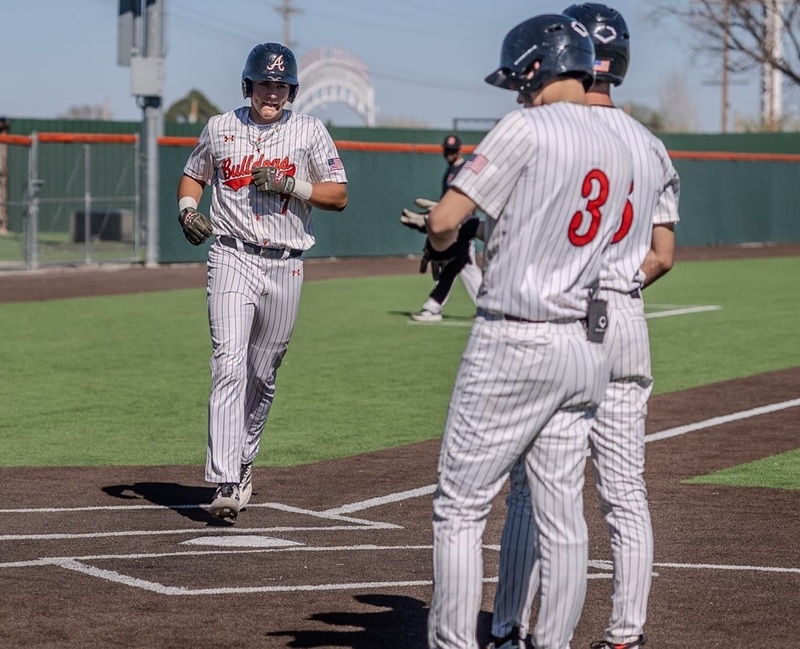 A teen boy in a grey and orange baseball uniform runs across home plate as two teammates watch.