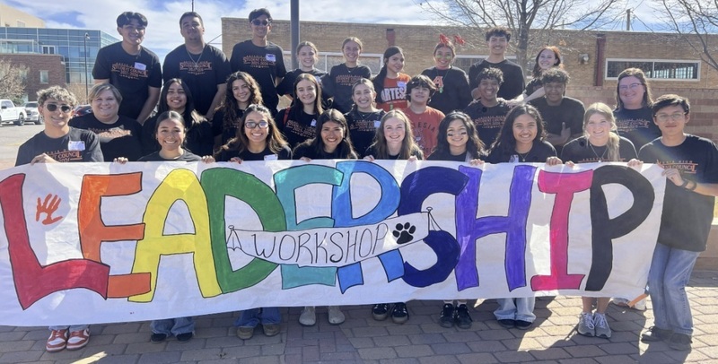 A large group of teens pose for a photo while holding a colorful banner that reads "Leadership Workshop."