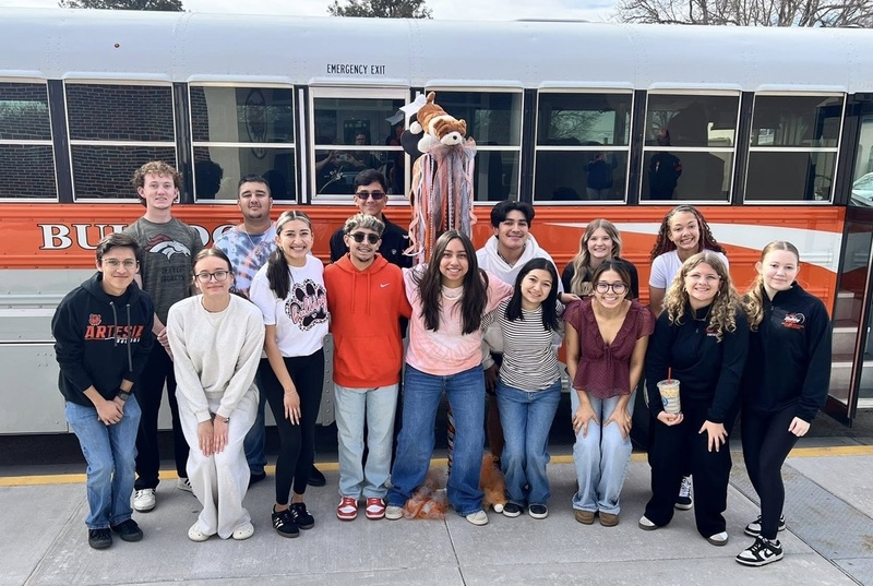 A group of teens pose for a photo outside a white and orange bus. One teen holds a large spirit stick with orange, white and black ribbons hanging from it and a stuffed Bulldog on top.