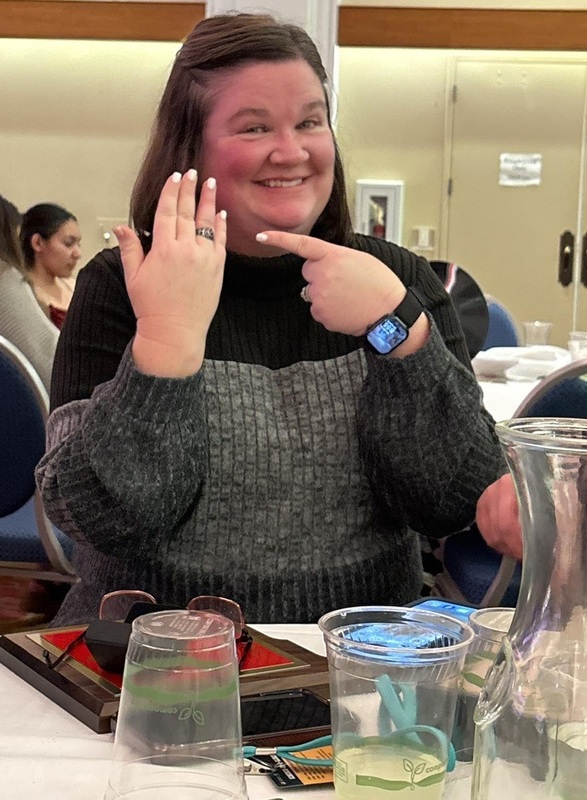 A woman smiles as she points to a class ring on her right hand. She is seated at a table with drinkware on it.