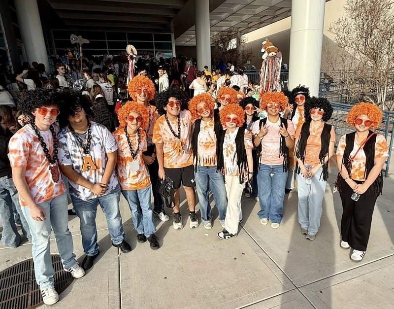 A group of teens pose for a photo. All are wearing orange tie-dyed shirts and jeans, round orange sunglasses, beads, and either a black or an orange afro wig. Some also wear black fringed vests.