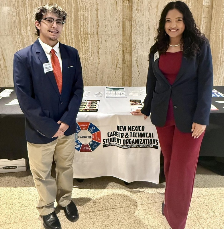A teen boy in a suit and a teen girl in a pantsuit and jacket smile for the camera while standing in front of a table reading "New Mexico Career and Technical Student Organizations."