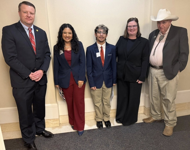 A teen boy in a suit and a teen girl in a pantsuit and jacket stand alongside a woman in a black outfit and two men in suits. One of the men wears a cowboy hat.