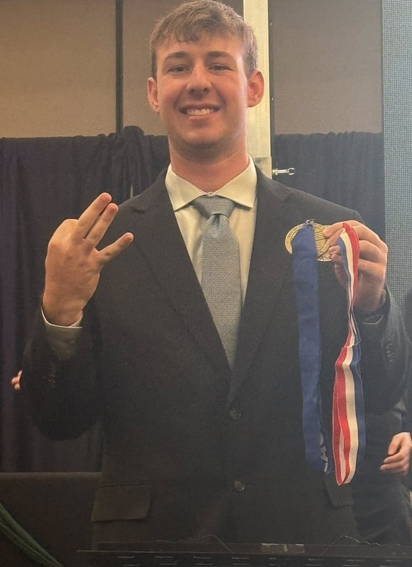 A teen boy in a suit smiles for the camera. Sith one hand, he holds up three fingers. With the other, he holds two medals.