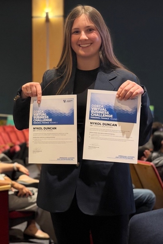 A teen girl in a suit smiles for the camera as she holds up two certificates.