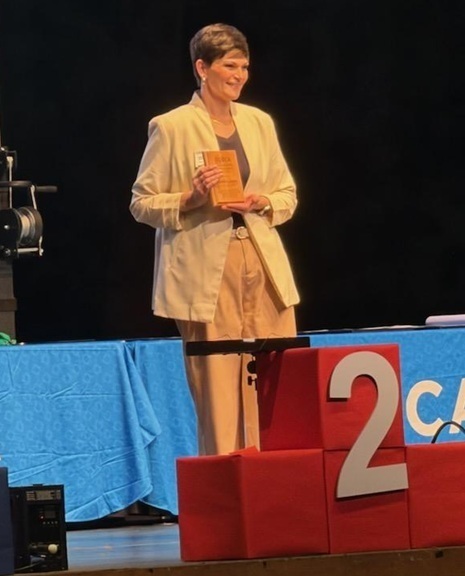 A woman in a light-colored pant suit and jacket holds a plaque as she smiles for the camera.
