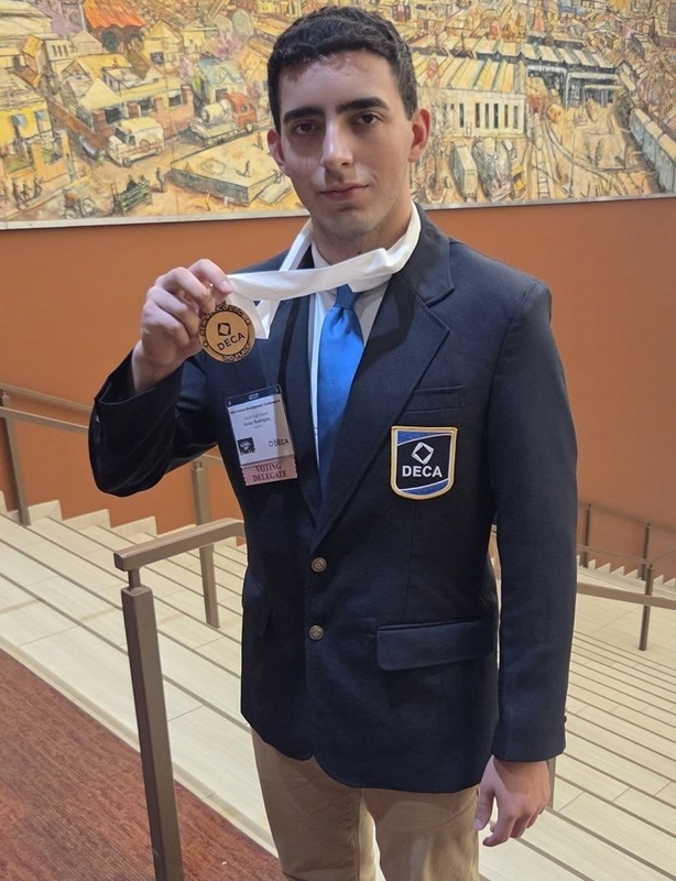A teen boy in a suit bearing a patch reading "DECA" poses for the camera. With his right hand, he holds up a bronze medal he is wearing around his neck.