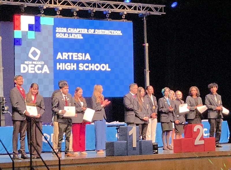 A group of teens stand in a line on a stage. Behind them, a large screen reads "2026 Chapter of Distinction, Gold Level: Artesia High School."