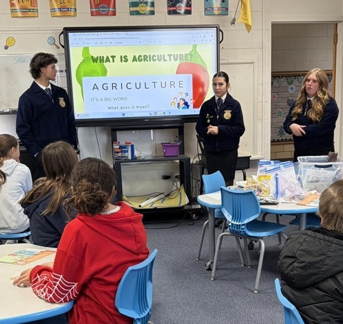 Three teens in blue FFA jackets stand alongside a screen at the front of an elementary school classroom as young students seated at tables watch them.
