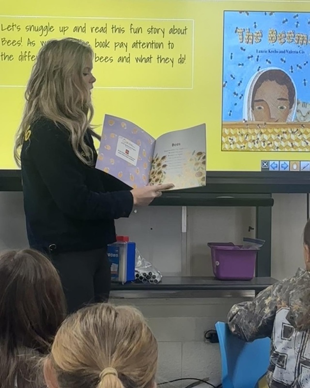 A teen girl in a blue FFA jacket holds a book open as she reads to young students in an elementary school classroom.