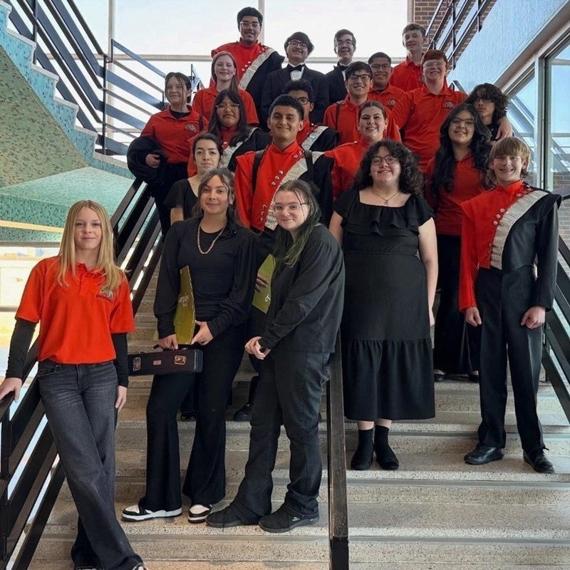 A group of teens, three in black semi-formal attire and the rest in orange band uniforms, pose for a photo on a set of stairs in an airy building foyer.