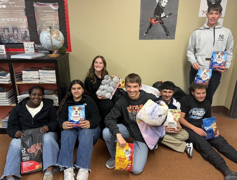 A group of teens, all but one seated on the floor, pose for a photo holding various articles of pet food and supplies.