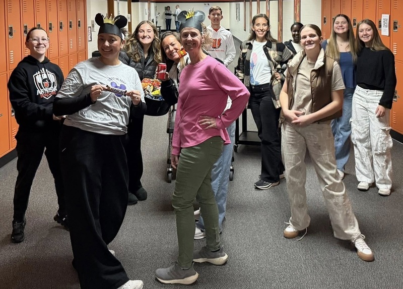 A group of teens and two female teachers pose for a photo in a school hallway. The teachers are retrieving items from a beverage and snack cart.