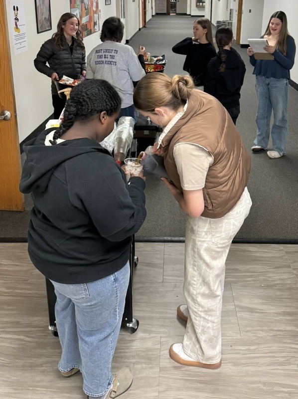 One female teen holds a cup of ice as another female teen pours a beverage over it.