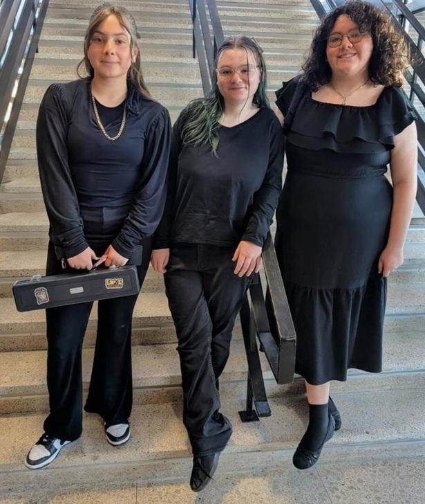 Three teen girls in semi-formal black attire pose for a photo at the bottom of a set of stairs. One girl holds a black instrument case in her hands.