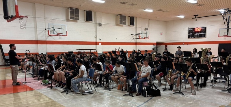 A large group of preteens holding various instruments and seated in chairs in a gymnasium look at a male band director at the front of the room.