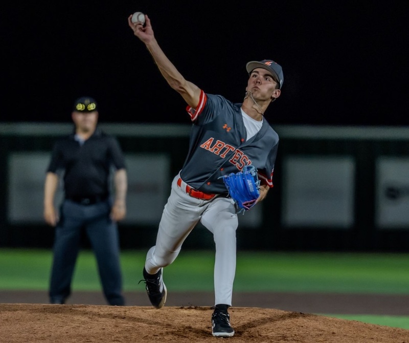 A teen boy in a grey and orange baseball uniform strains as he prepares to release the ball from his right hand on the mound. He is wearing a blue glove on his left hand.