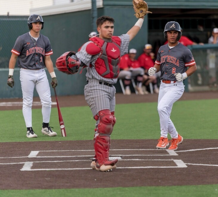 A teen boy in a grey and orange baseball uniform prepares to step on home plate as a teammate in the background watches and an opposing catcher in red and grey holds his glove hand above his head and looks out into the field.