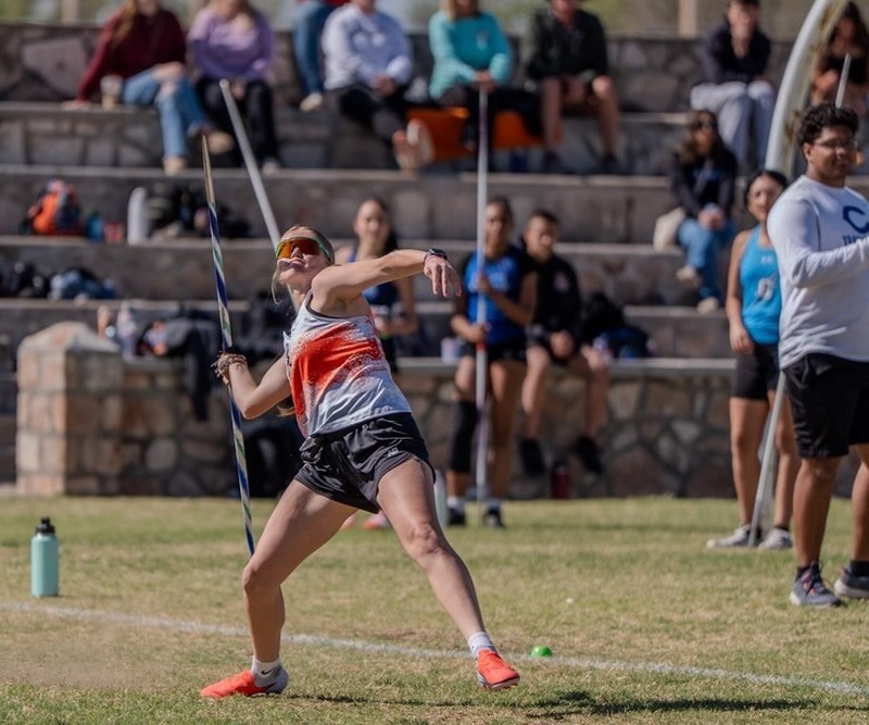 A teen girl in a grey and orange track uniform and orange-tinted sunglasses rares back as she prepares to throw a javelin. 