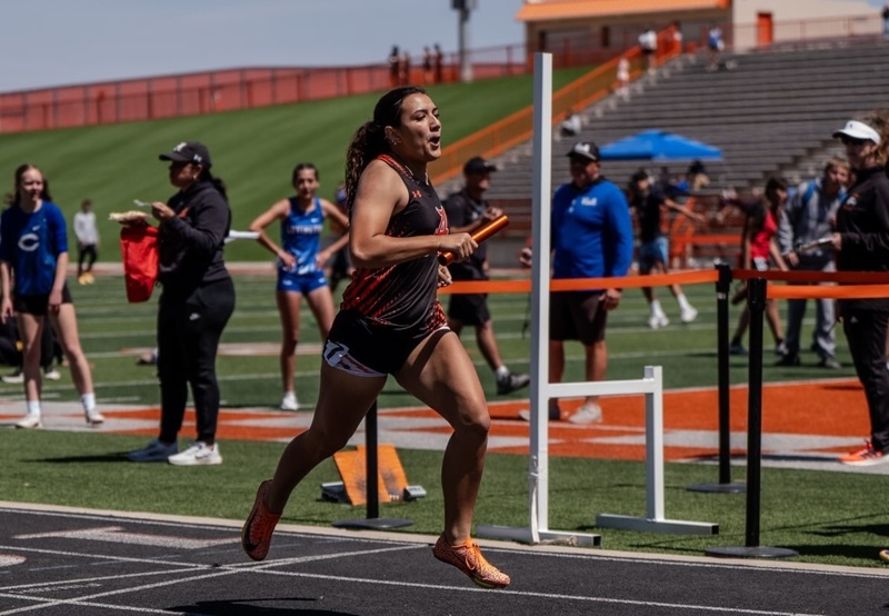 A teen girl in a black and orange track uniform runs along a track past timers standing on a busy infield.