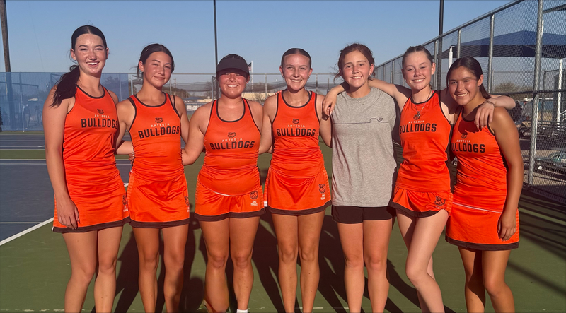 Seven teen girls, six in orange tennis uniforms and one in a grey top and black shorts, stand in a line with their arms around each other and pose for a photo on a tennis court.