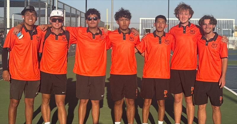 Seven teen boys in orange and black tennis uniforms smile for the camera with their arms around each other as they pose for a photo on a tennis court.