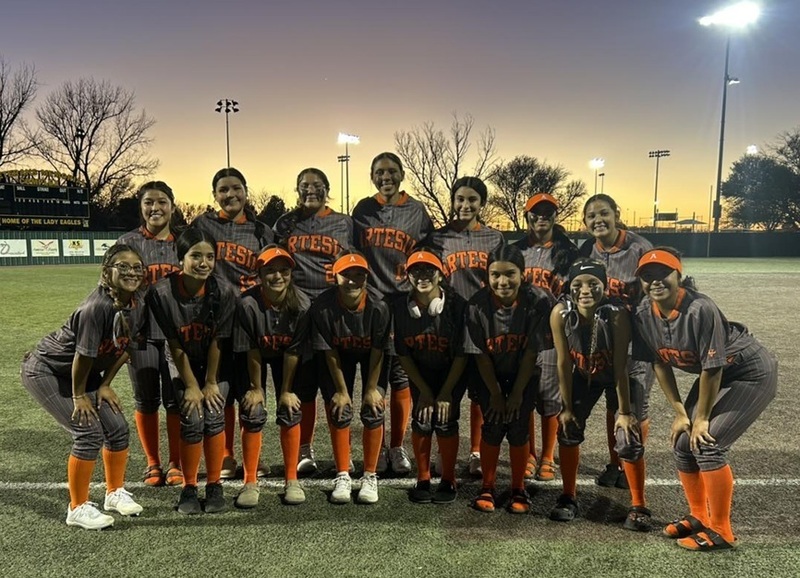 A high school softball team in grey and orange uniforms poses for a photo on a field with a sunset behind them.