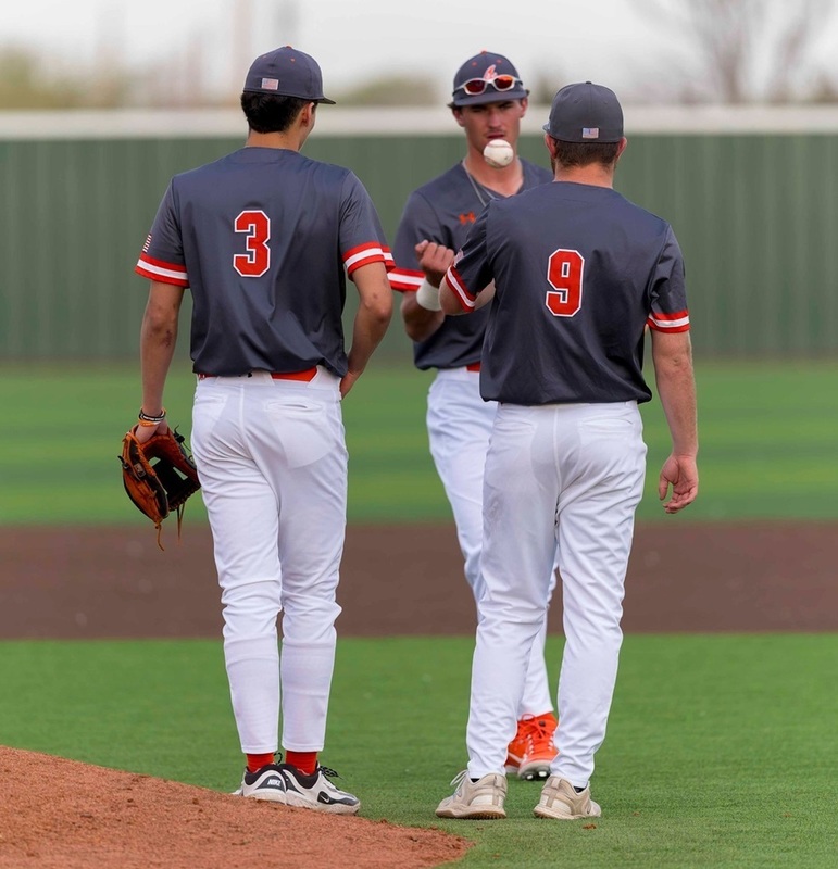 Three teen boys in grey, orange and white baseball uniforms stand near the mound as one tosses the ball to another.
