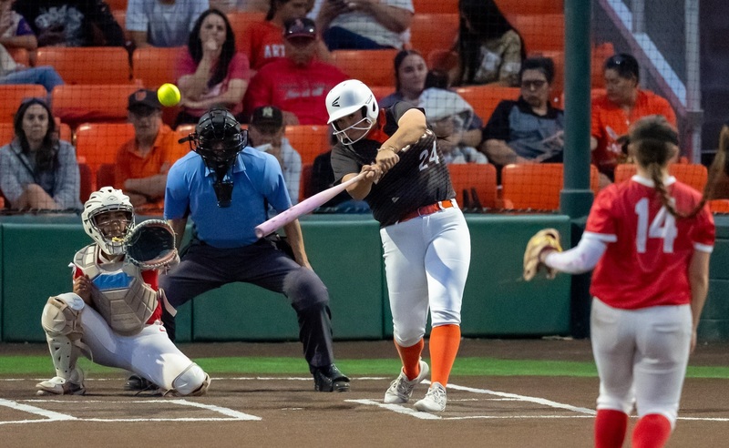 A teen girl in a black, orange and white softball uniform launches the ball off the bat as the opposing catcher and pitcher, in red and white, and fans in the stands behind home plate look on.
