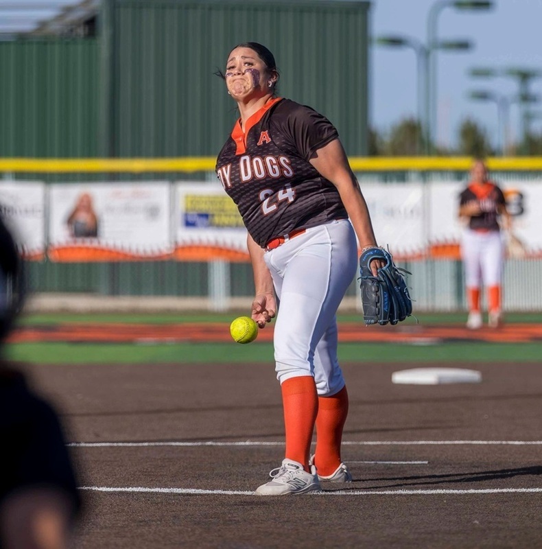 A teen girl in a black, orange and white softball uniforms releases the ball from her right hand in the pitcher's circle.