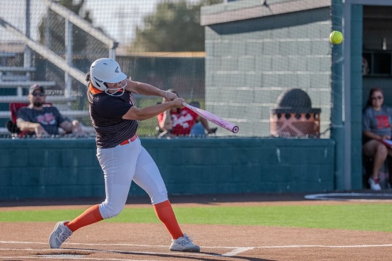A teen girl in a black, orange and white softball uniform swings as the ball flies off her bat into the air.