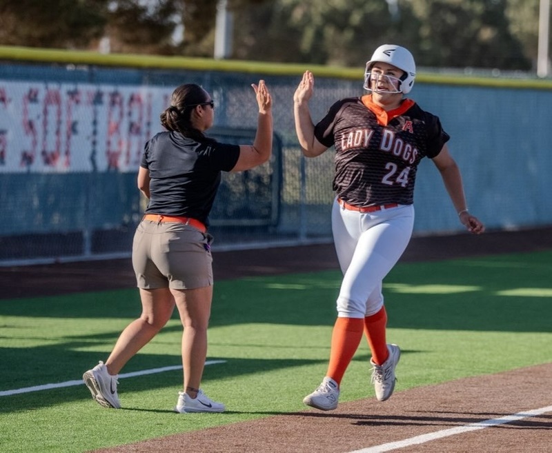 A teen girl in a black, orange and white softball uniform prepares to high-five a female coach in a black top and khaki shorts as she rounds third base.