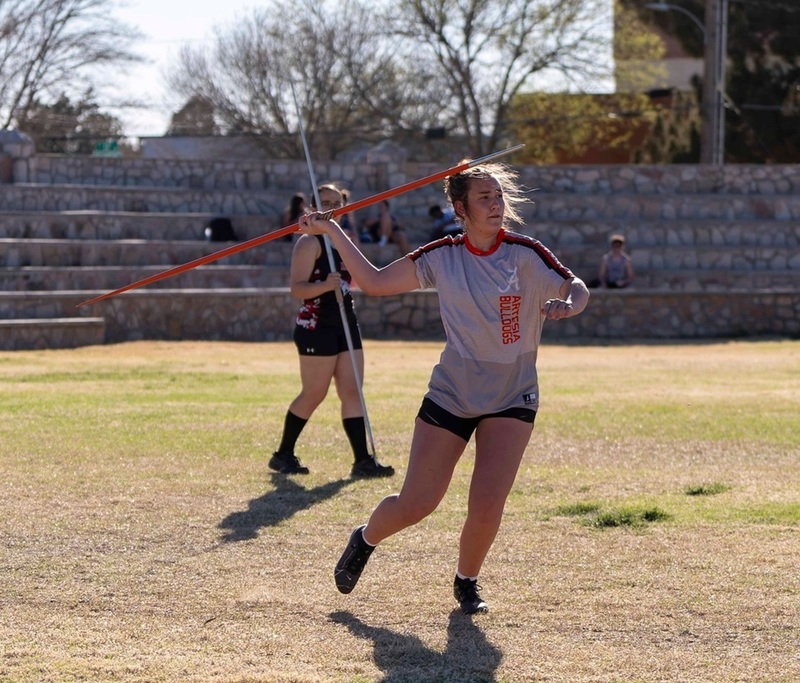 A teen girl in a grey and orange track uniform prepares to throw a javelin with her right hand.