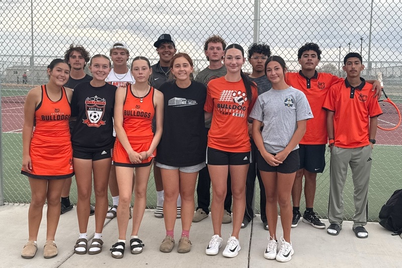 A group of male and female teens in orange, black and grey tennis uniforms pose for a photo outside a tennis complex.