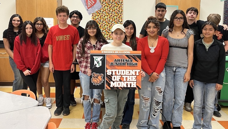 A teen girl surrounded by other teens holds a sign that reads "Student of the Month".