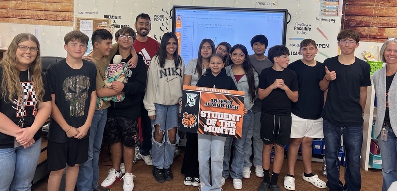 A teen girl surrounded by other teens holds a sign that reads "Student of the Month".