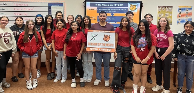 A male teacher surrounded by teen students holds a sign that reads "Teacher of the Month".