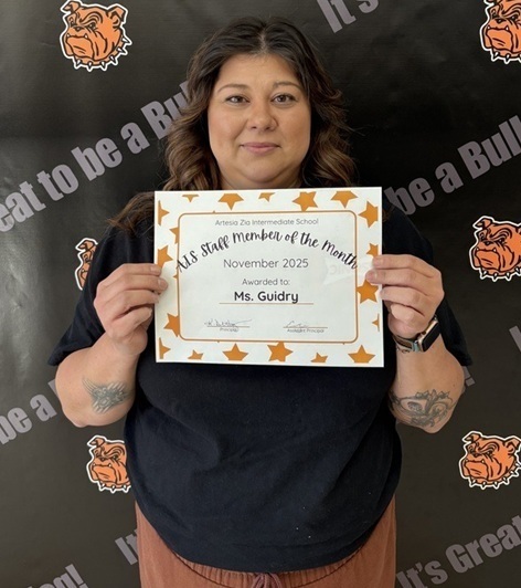 A female teacher in a black top holds a certificate that reads "Teacher of the Month".