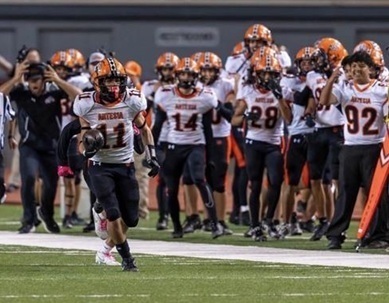 A teen boy in a white, orange and black football uniform runs up the field carrying the ball in his right arm as teammates cheer from the sideline.
