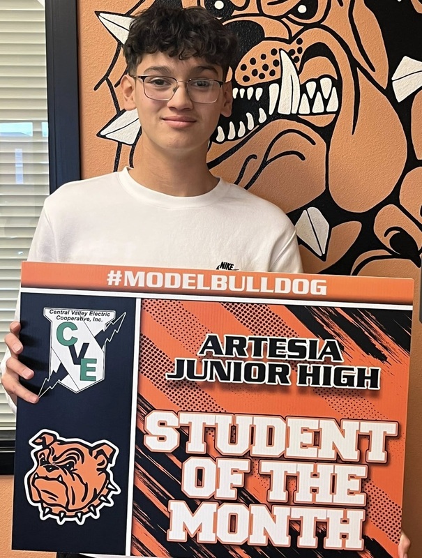 A teen boy in a white shirt, black hat and glasses holds a sign reading "Student of the Month".