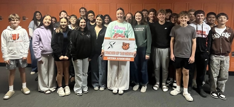 A female teacher surrounded by teen students holds a sign reading "Teacher of the Month".