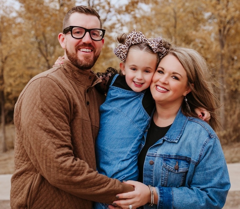 A female teacher in a jean jacket poses alongside her husband and young daughter.