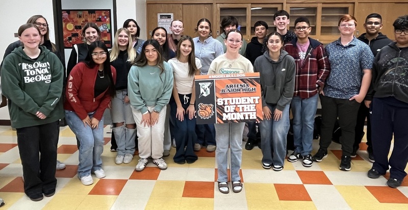 A teen girl surrounded by other teen students holds a sign reading "Student of the Month".