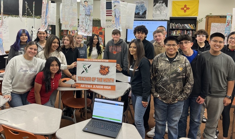 Teen students in a classroom hold a sign reading "Teacher of the Month".
