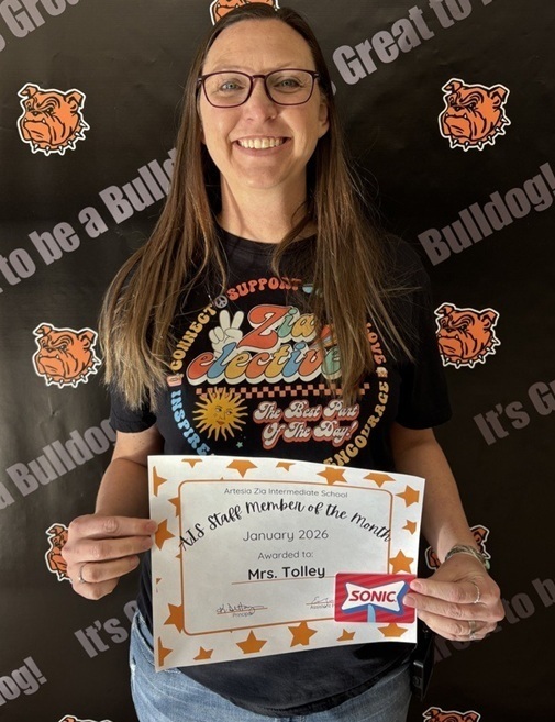 A female teacher in a black shirt holds a certificate reading "Teacher of the Month".