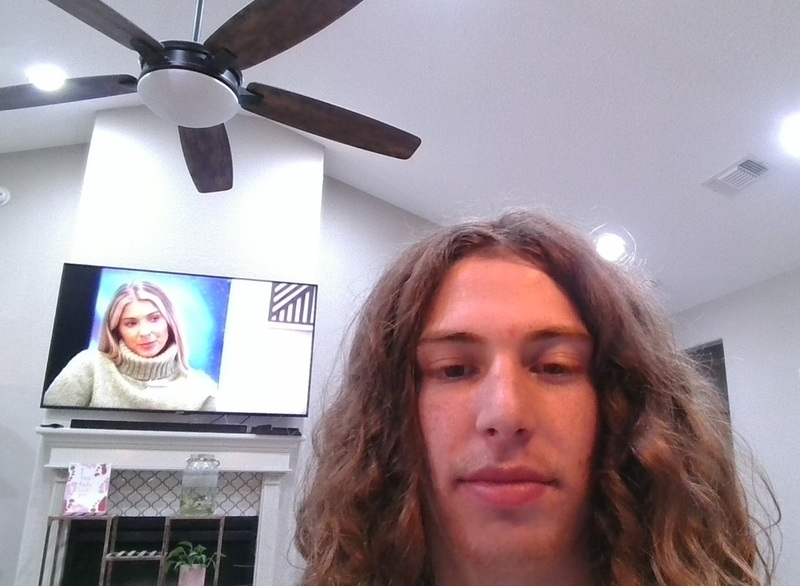 A long-haired teen boy poses for a photo in a living room in front of a TV with a woman on the screen.