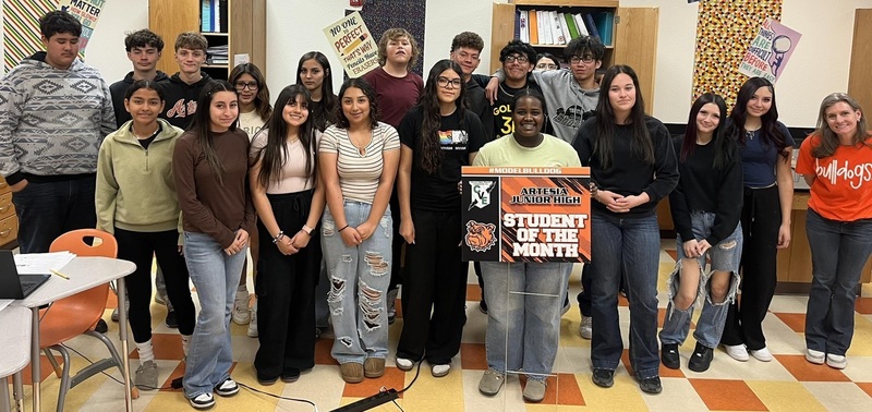 A teen girl surrounded by other teens holds a sign reading "Student of the Month".