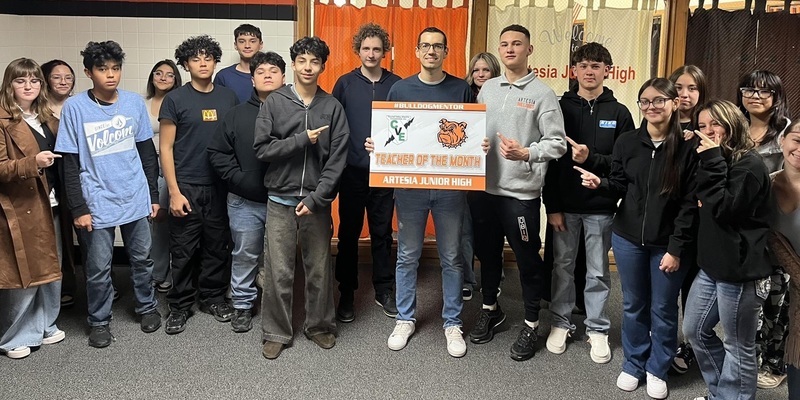 A male teacher surrounded by teen students holds a sign reading "Teacher of the Month".
