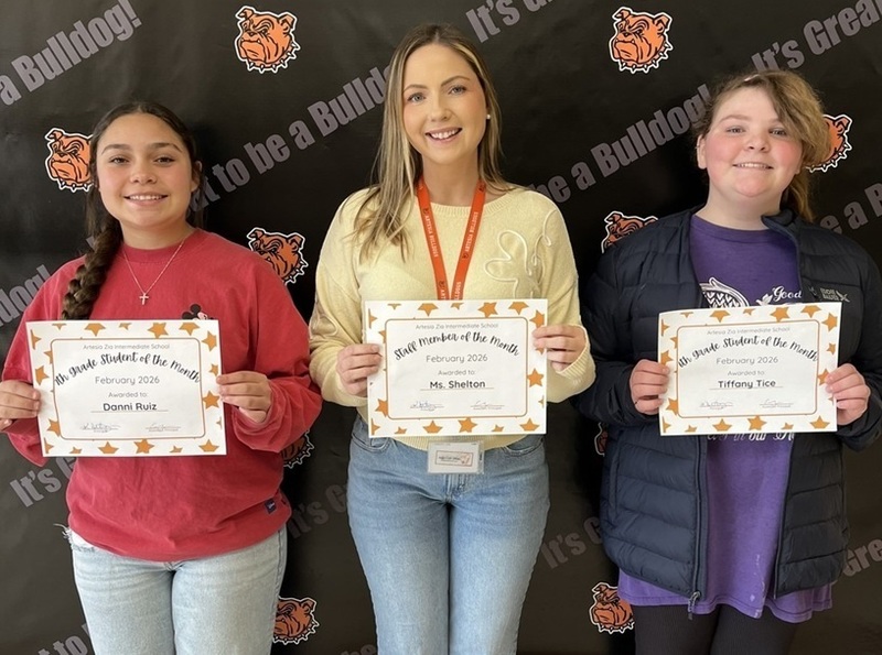 A female teacher in a yellow top stands between two preteen female students. All hold certificates.